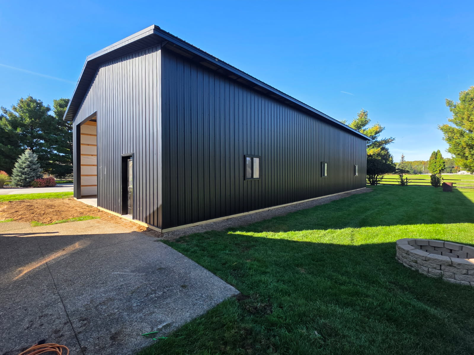 Large black metal shop pole barn with three side windows, concrete walkway, and fire pit in a Northern Indiana backyard
